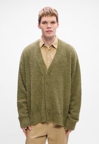 Groene textuur cardigan met een losse pasvorm, v-hals, voor knopen en geribde manchetten, gelaagd over een lichtbruine overhemd met kraag.