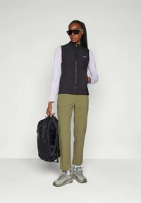 Mulher a usar um colete preto, camisa de manga longa clara, calças oliva, sapatilhas cinzentas, óculos de sol pretos, segurando uma mochila preta ao lado.