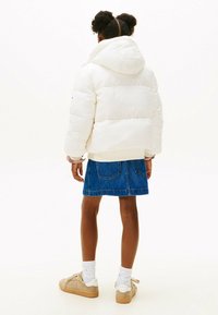 Witte pufferjack met een hoge kraag en capuchon, gecombineerd met een blauwe denim rok. Model draagt lichte schoenen en witte sokken.