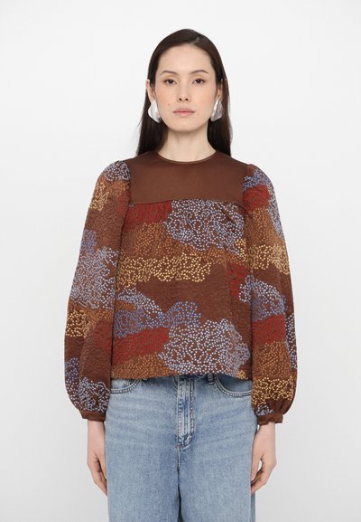Blouse marron à motifs avec des manches ballon texturées, affichant des motifs bleu, rouge et or. Portée avec des jeans en denim bleu clair.