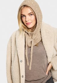 Beige plüss kapucnis pulóver bordás textúrával, amelyet egy krémszínű, bolyhos külsejű kabát és egy barna alapréteg egészít ki.