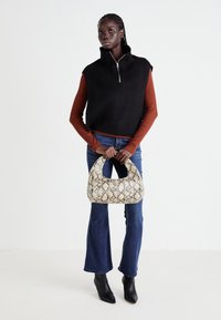 Frau steht und hält eine beigere Handtasche im Schlangenledermuster, trägt einen schwarzen ärmellosen Strickpullover über einem rostfarbenen Langarmshirt, blaue Jeans und schwarze Stiefel.