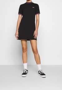 Robe t-shirt en coton noir à manches courtes, avec un bord à carreaux roses et un logo brodé sur la poitrine. Associée à des baskets noires.