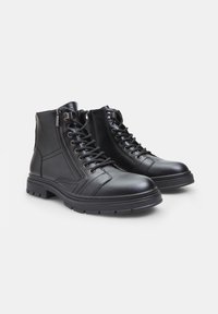 Botas de tobillo de cuero negro con un diseño de cordones en la parte delantera y cremalleras laterales. Suela de goma gruesa, acabado texturizado y detalles cosidos.