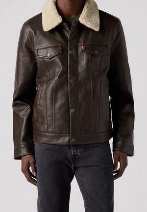 Homme portant une veste en cuir marron foncé avec un col en shearling crème et un jean foncé, debout devant un fond clair uni.