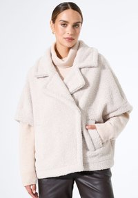 Beige, flauschige Fleecejacke mit breitem Kragen, kurzen Ärmeln und einer Fronttasche; darüber trägt sie einen hellbeigen Rollkragenpullover.