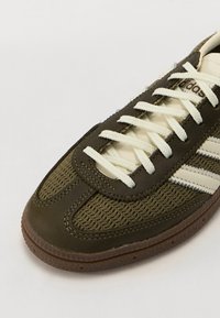 Olivgrön sneaker med en texturerad tygöverdel, som har vita snörningar, en tanfärgad gummisula och tre vita ränder för varumärket.