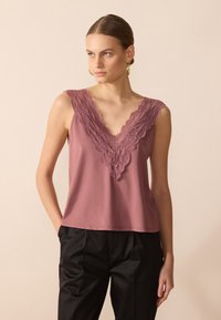 Top de tirantes color rosa con un amplio escote en V de encaje. Tejido suave y liso; corte holgado; combinado con pantalones negros. El detalle de encaje añade textura.