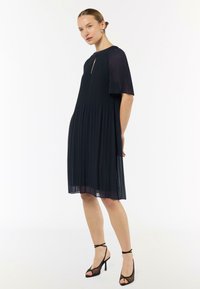 Czarna plisowana sukienka z krótkimi rękawami, z wycięciem w kształcie łezki przy szyi oraz warstwowym, zwiewnym krojem; zestawiona z czarnymi sandałami na obcasie.