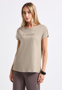 Beige tričko s krátkým rukávem a kulatým výstřihem, na přední straně je potisk "CANNES". Hladká textura, volný střih, kombinováno s tmavými kalhotami.