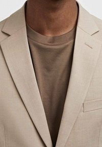 Blazer beige con rever a punta sopra una maglietta girocollo marrone. Tessuto liscio, dettagli minimali e accento sulla tasca discreto.