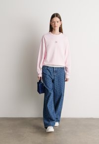 Różowa bluza z małym graficznym logo, zestawiona z luźnymi, niebieskimi spodniami w wzory i białymi trampkami. Trzyma małą niebieską torebkę.