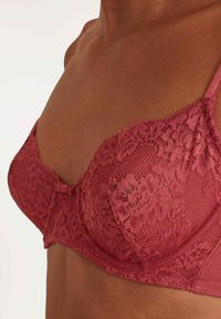Kant BH in dieproze kleur met doorschijnende en gestructureerde stof. Kenmerken omvatten bloemmotieven, verstelbare bandjes en een centrale strikaccent.