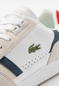 Lacoste-sneaker med ett vitt och beige läderövre, marinblå accenter, perforerade detaljer och en grön broderad krokodil-logotyp.