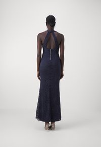 Abito da sera in pizzo navy con silhouette aderente, collo halter, schiena scoperta e chiusura con zip. Presenta un motivo floreale e orlo lungo fino al pavimento.