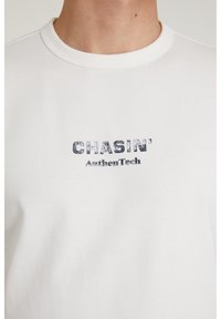 Bijela pamučna majica s plavim istrošenim natpisom "CHASIN' AuthenTech" na prsima, okrugli izrez i kratki rukavi.