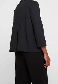 Veste noire courte à coupe ample, manches trois-quarts, col large, avec un pli central dans le dos pour plus de liberté de mouvement.