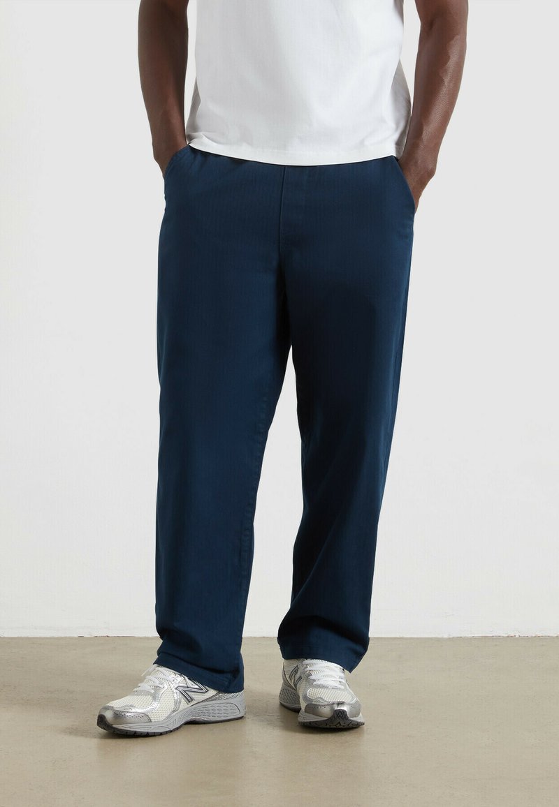 Granatowe spodnie o gładkiej fakturze i elastycznym pasku, z kieszeniami po bokach, zestawione z srebrnymi butami sportowymi.