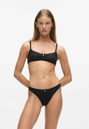 Femme aux cheveux longs portant un ensemble de lingerie noir deux pièces avec de petits nœuds sur le soutien-gorge et la culotte, sur un fond blanc uni.
