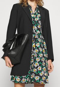 Schwarzer Blazer über einem floralen Kleid mit einer schwarzen Handtasche. Das Kleid zeigt Gänseblümchen in Weiß, Gelb und Grün auf einem schwarzen Hintergrund.