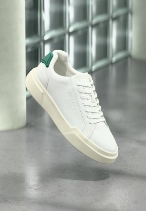 Hvid lavtop-sneaker med grøn hælkappe, tyk off-white sål og præget brandnavn, vist mod en reflekterende glasblokvæg.