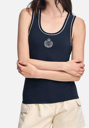Donna che indossa un top senza maniche blu navy con rifiniture bianche ed emblema, abbinato a pantaloncini beige, braccia incrociate sul torso.