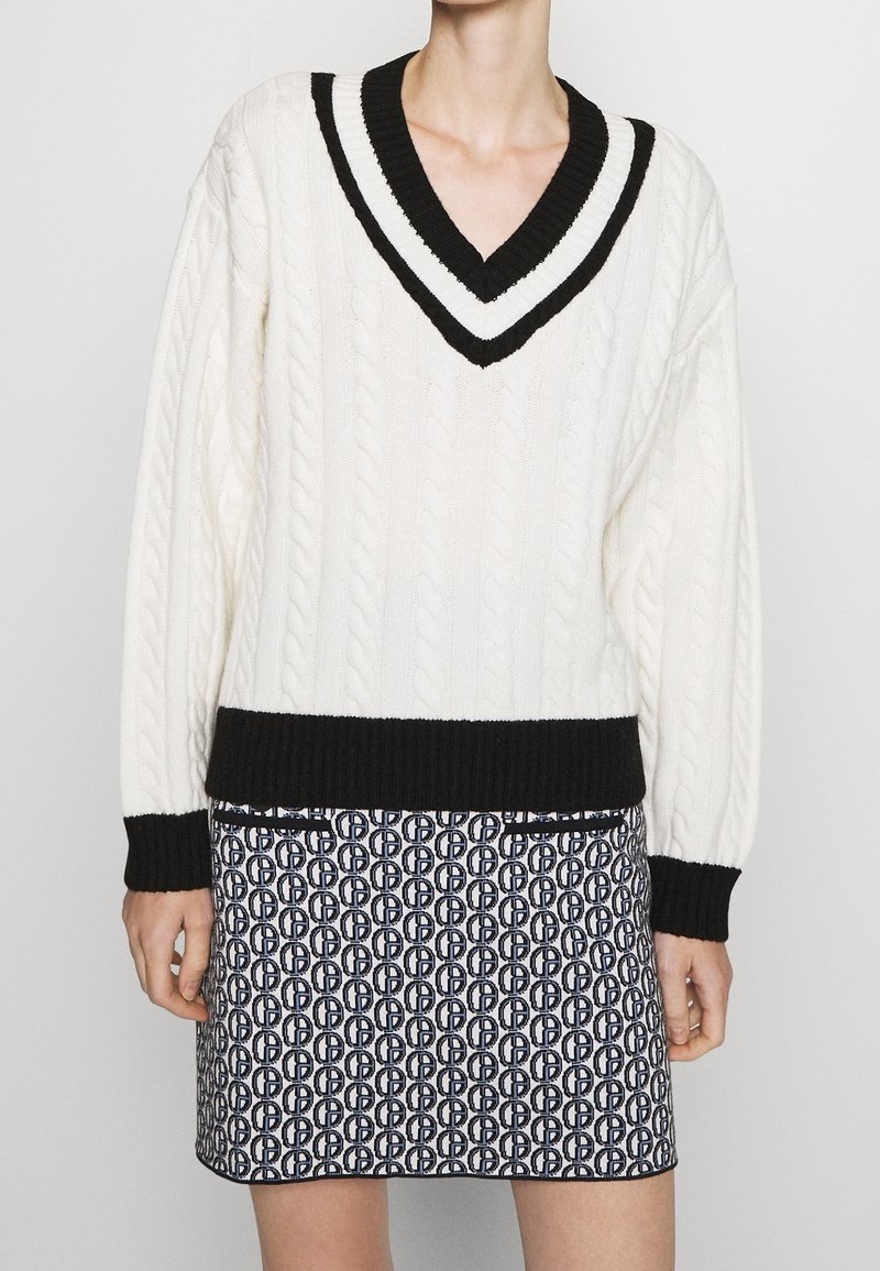 Bílý svetr s copánkovým vzorem, černým výstřihem ve tvaru V a lemy rukávů, doplněný vzorovanou sukní s opakujícím se kruhovým motivem v námořnické modři a bílé barvě.