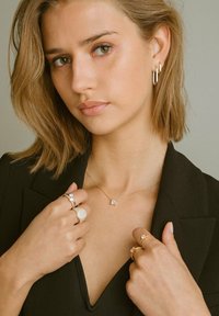 Guldhalsband med fyrkantigt hänge, flera ringar och örhängen; modell iklädd en svart blazer, mjuk textur, och neutral bakgrund.