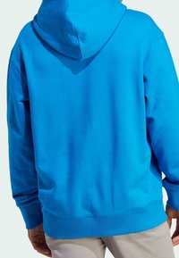 Man med ljusblå hoodie och ljusa byxor, sedd bakifrån mot en enfärgad ljus bakgrund.