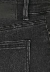 Mustat farkut, joissa on tekstuuripinta, takatasku ja nahkalamppu, johon on kaiverrettu "PIECES". Ompeleet saumoissa.