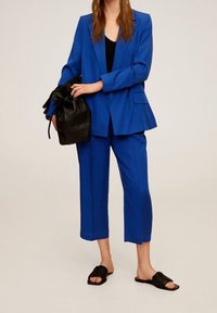Costume bleu sur mesure avec un blazer ample et un pantalon court, assorti à des sandales noires à enfiler et un sac à cordon noir. Texture de tissu lisse.