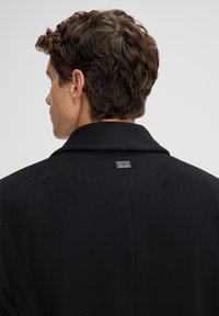 Cappotto in lana nera con colletto ampio, texture liscia e una piccola etichetta con logo sul retro. Mostra la vista posteriore del capo.