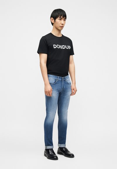 Άνδρας που στέκεται μπροστά σε λευκό φόντο, φορώντας μαύρο μπλουζάκι DONUP, ξεθωριασμένα μπλε τζιν με ρεβέρ και μαύρα δερμάτινα παπούτσια.