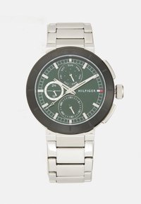 Montre HILFIGER avec un cadran analogique vert, un bracelet en acier inoxydable argenté, une lunette noire, des sous-cadrans pour le jour et la date, et une couronne texturée.