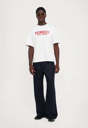 Mann trägt ein weißes Fiorucci Cash & Carry T-Shirt, dunkle weite Hose, schwarze Schuhe und rechteckige Sonnenbrille, steht vor einem einfarbigen Hintergrund.