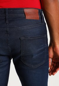 Dark blue denim jeans with a smooth texture. Features a leather patch with brand logo at the back waist and standard back pocket design.