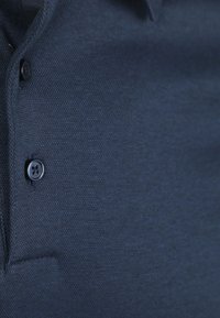 Close-up van een donkerblauwe textuurstof shirt met twee zwarte knopen en een deel van een zak.