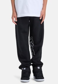 Schwarze Jogginghose mit lockerem Schnitt und weißer Markenkennzeichnung an der Seite. Kombiniert mit schwarzen High-Top-Sneakern und einem weißen T-Shirt.