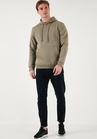 Olivovozelená mikina s nastaviteľnými šnúrkami, klokankou a rebrovaným lemom, v kombinácii s tmavými slim fit nohavicami a čiernymi teniskami.