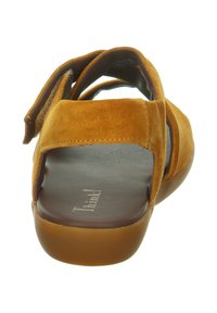 Braune Wildledersandale mit offenem Rücken, verstellbarem Riemen und einer glatten, dunklen Innensohle. Die Sohle ist flexibel und leicht durchscheinend.