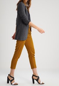 Cárdigan gris de manga larga, pantalones cortos mostaza y sandalias negras de tacón con acentos blancos. Tela suave y corte entallado.