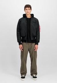 Zwarte gewatteerde bomberjack met een capuchon, ritsluiting, ribgebreide manchetten en een logo patch. Gemaakt over een zwart shirt en olijfgroene broek.