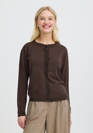Femme blonde souriante portant un cardigan marron foncé boutonné et un pantalon à carreaux beige sur un fond blanc uni.