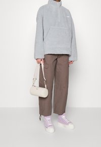 Helles grauer Fleece-Pullover mit einem halben Reißverschluss, Fronttasche; braune weite Cargo-Hosen; lavendelfarbene Socken; cremefarbene zylindrische Tasche und weiße Sneakers.