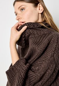 Ciemnobrązowy sweter wykonany na drutach z wysokim kołnierzem i teksturowanym wzorem, z grubymi włóknami i luźnym krojem, trzymany przez rękę przy kołnierzu.