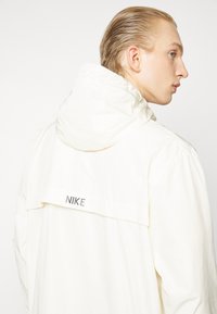 Hvid Nike-jakke med hætte, der har et logo bagpå. Materialet ser glat ud med et let design og en afslappet pasform.