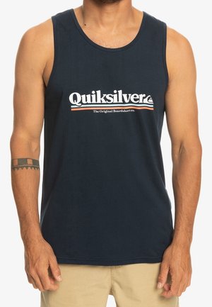 Mężczyzna w granatowym bezrękawniku Quiksilver i beżowych szortach, z czarnym tatuażem w stylu tribal na lewej przedramieniu.