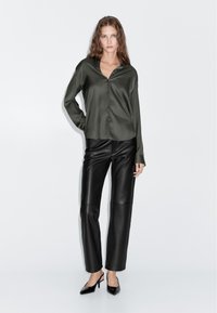 Donkergroene satijnen blouse met knopen en een puntige kraag, gecombineerd met zwarte, hooggesneden leren broek en zwarte stiletto's.