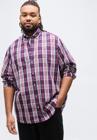 Άνδρας με dreadlocks που φοράει πουκάμισο καρό κόκκινου, λευκού και μπλε χρώματος, με τα μανίκια ανασηκωμένα και το ένα χέρι στην τσέπη, μπροστά από απλό φόντο.