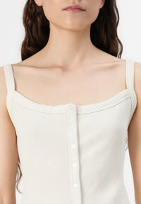 Cream ribbed tank top with a square neckline, thin straps, and a row of five buttons down the front. Soft texture, fitted design.